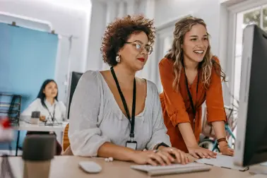 Zwei Frauen schauen in einem Büro lächelnd zusammen auf einen Bildschirm. 